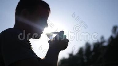一个年轻健康的人从玻璃杯中喝干净的水，站在轮廓、<strong>光线</strong>和<strong>光线</strong>中的太阳旁边。